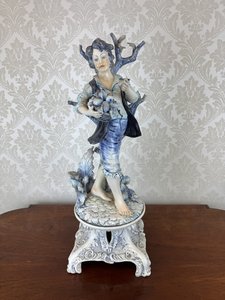 Porcelánová figurka mladíka s ovocem u stromu na podstavci s modrobílou patinou